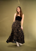 Load image into Gallery viewer, A person wearing a long, floral Coyote skirt with a high-low hemline and an adjustable waist, paired with a black top and white shoes, standing against a yellow background.