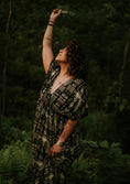 Load image into Gallery viewer, Woman in a patterned dress standing in a forest, holding an object above her head.
