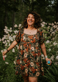 Load image into Gallery viewer, Woman in a floral dress standing in a garden with flowers and greenery.