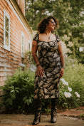Load image into Gallery viewer, Woman in a patterned dress standing outdoors near a New England style house with greenery.