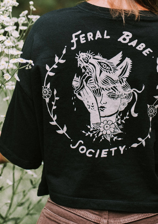 Black t-shirt with 'Feral Babe Society' design on the back, worn by a person in a natural setting.