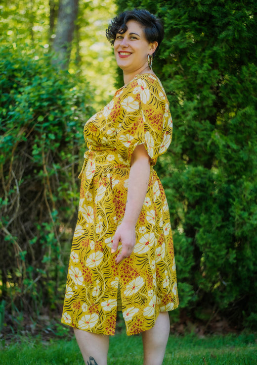 Woman wearing a yellow floral dress standing outdoors with greenery in the background